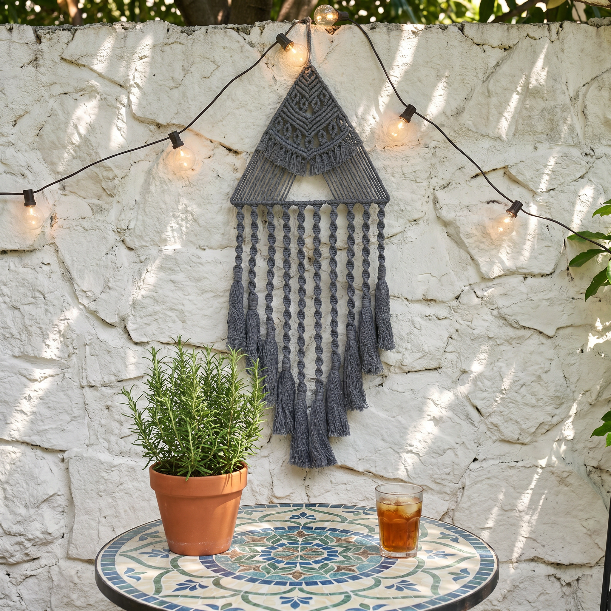 Gray triangle macrame dreamcatcher on outdoor stone wall with string lights — boho patio wall decor with cotton fringe