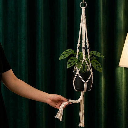 Hand holding tassel fringe of classic macrame plant hanger against dark emerald green curtain, boho decor detail