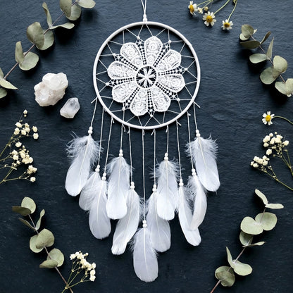 White dreamcatcher with feathers on a dark background with greenery and crystals