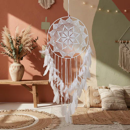 White dreamcatcher hanging in a cozy room with a wooden bench, vase, and pillows.