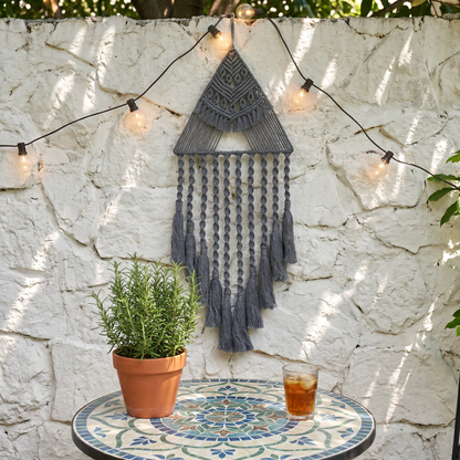Gray triangle macrame dreamcatcher on outdoor stone wall with string lights — boho patio wall decor with cotton fringe